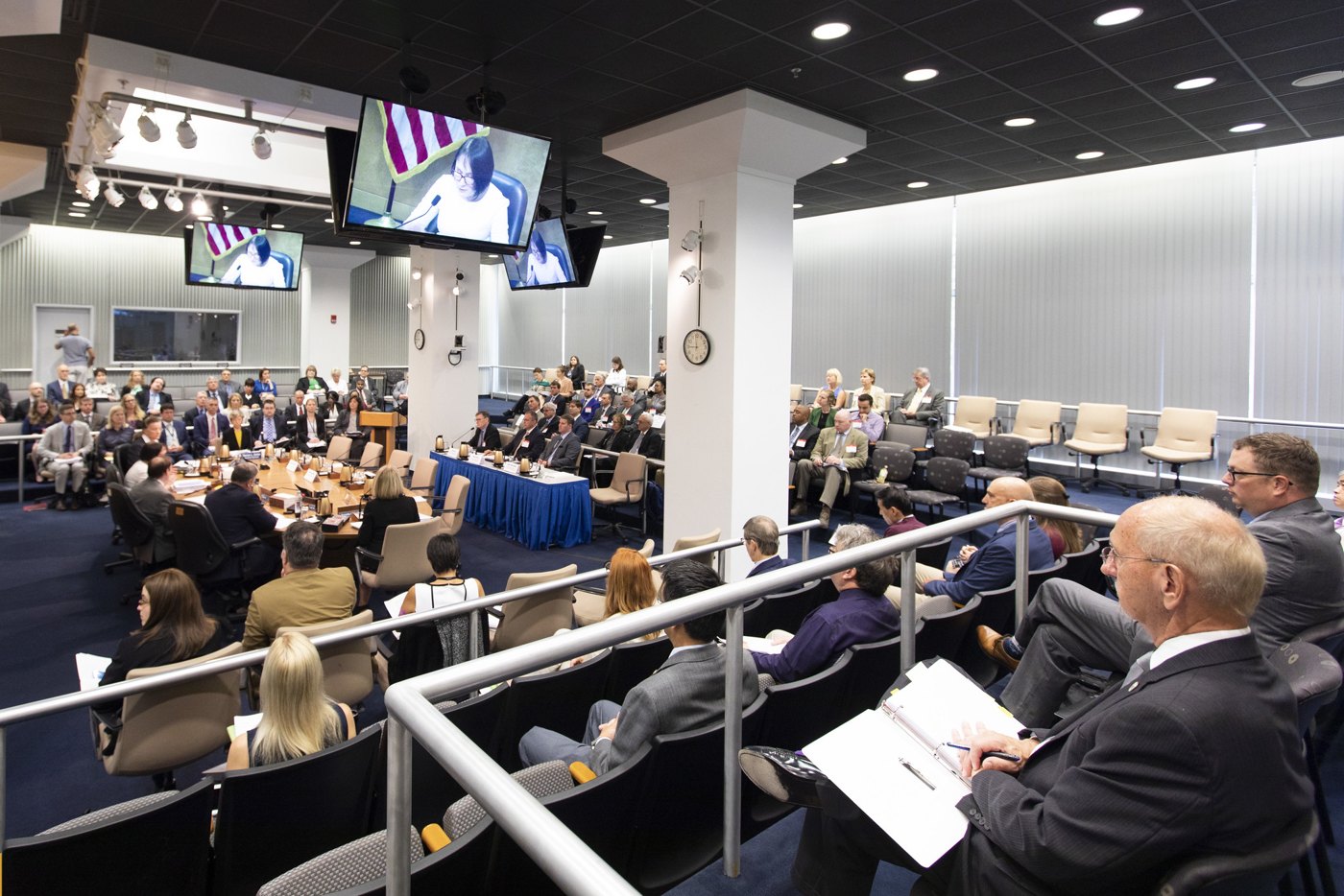 A full NRC hearing room seen from the public gallery, the commission at the table and the proceedings carried on overhead screens